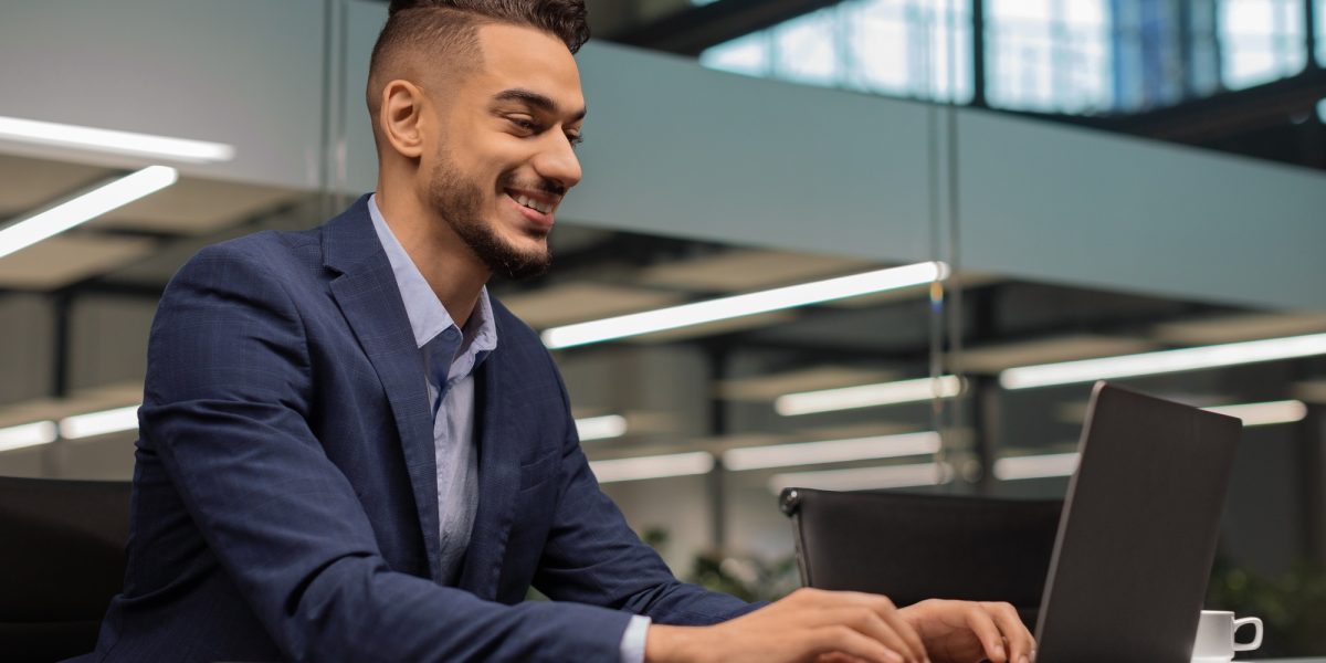 Cheerful middle eastern businessman typing on notebook, office interior